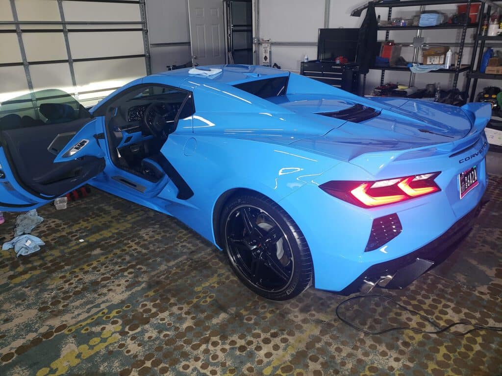 A bright blue Chevrolet Corvette sports car with its driver-side door open is parked in a garage with shelves, tools, and various items visible in the background. window tinting