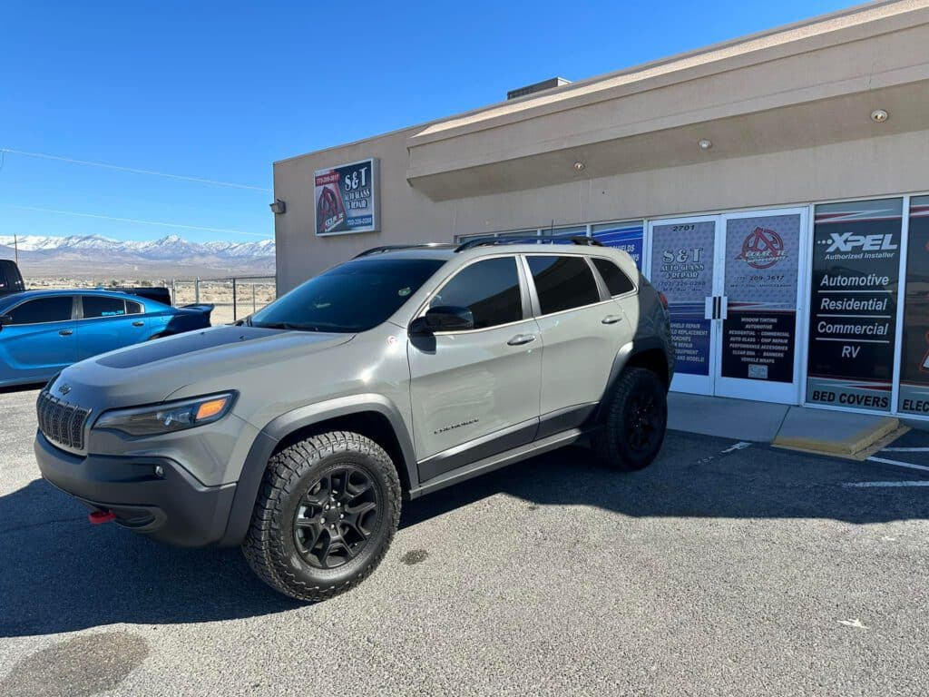 A gray Jeep Cherokee SUV with black wheels is parked in front of a detailing and automotive services shop on a sunny day, with mountains visible in the background. window tinting