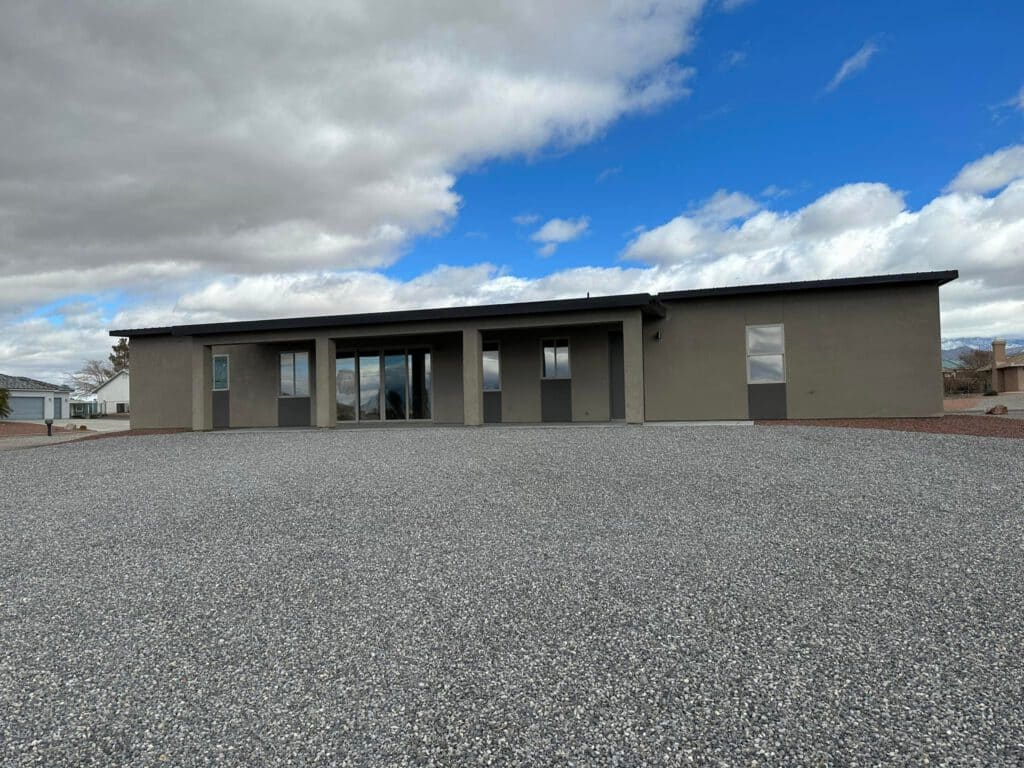 A modern, single-story house with large windows and a flat roof sits under a partly cloudy sky. The front yard is covered in gravel, and the house exterior is painted in neutral tones. window tinting