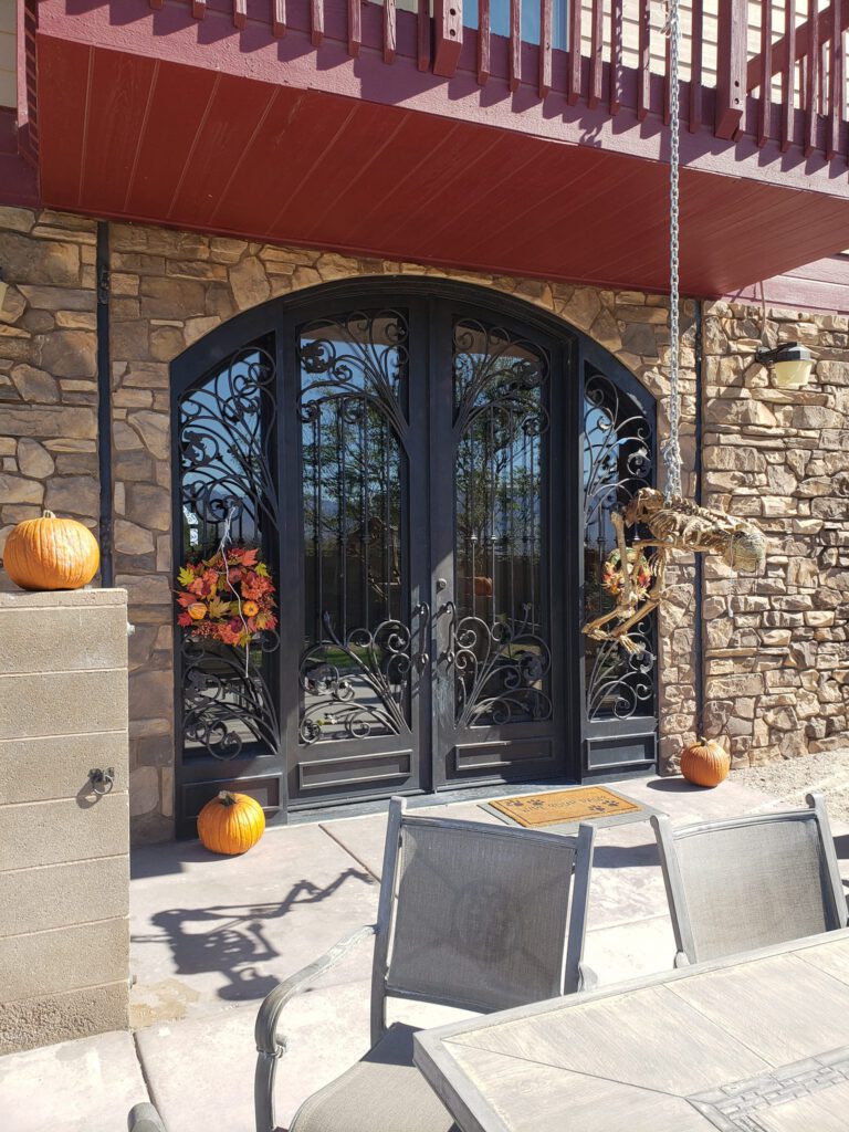 A black wrought iron double door with decorative designs is set in a stone wall. Pumpkins and a skeleton decoration hang nearby, along with autumn leaf wreaths on the doors. Patio chairs and a table are in the foreground. window tinting