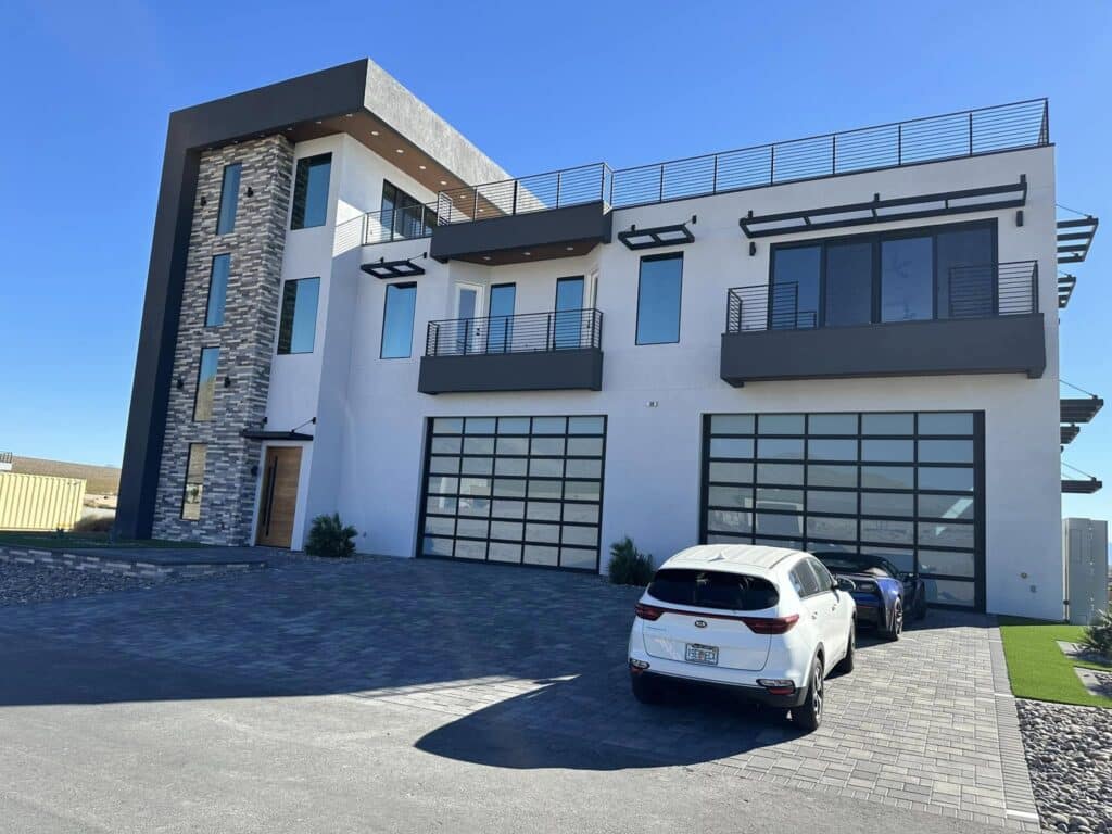 Modern three-story house with large glass garage doors, stone accent wall, multiple balconies, and two cars parked in the driveway on a sunny day. window tinting