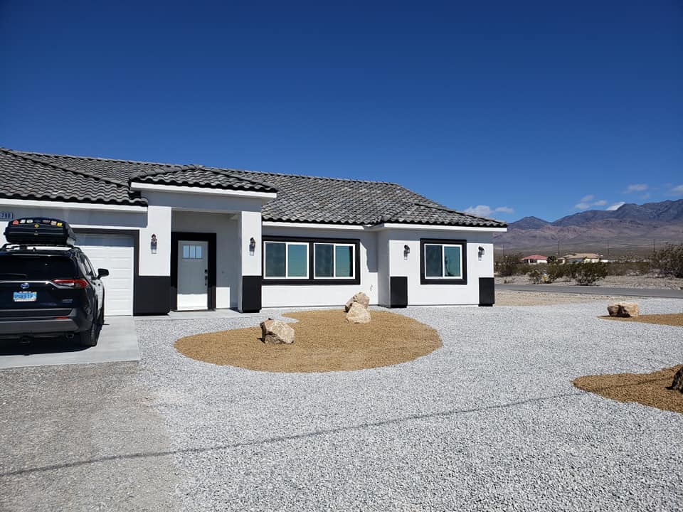 A white, single-story house with a gray tile roof sits on a gravel yard with circular areas of tan mulch and rocks. A black car is parked in the driveway. Mountains are visible in the background under a clear blue sky. window tinting